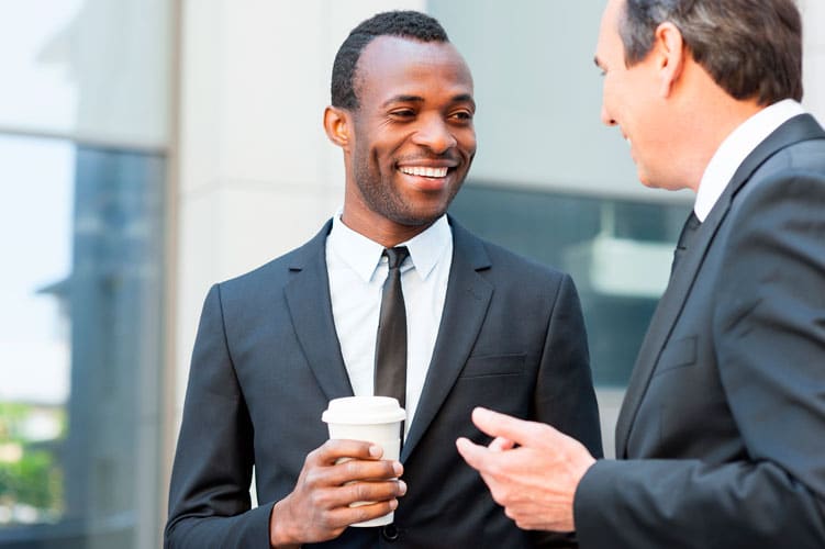 Two men in business suits are talking outside a modern office building. One man is smiling and holding a disposable coffee cup, while the other gestures as they discuss jobs for newcomers in Toronto.