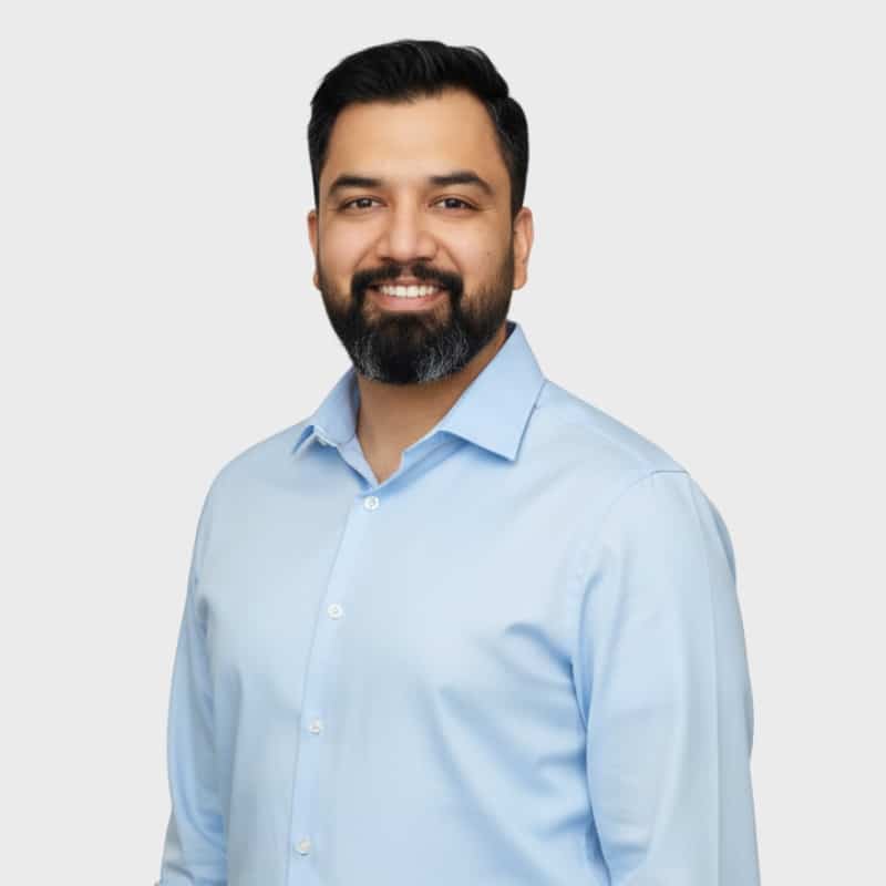 A man with dark hair and a beard smiles at the camera. He is wearing a light blue button-up shirt and stands against a plain, light gray background, representing an IT staffing agency in Toronto.